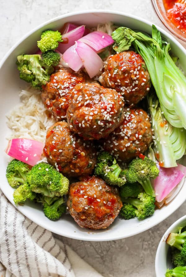 A bowl of rice topped with meatballs glazed in a red sauce, garnished with sesame seeds, and accompanied by broccoli, bok choy, and red onion.