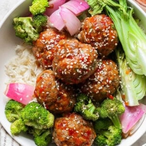 A bowl contains rice, meatballs with sauce and sesame seeds, broccoli, red onion, and bok choy.