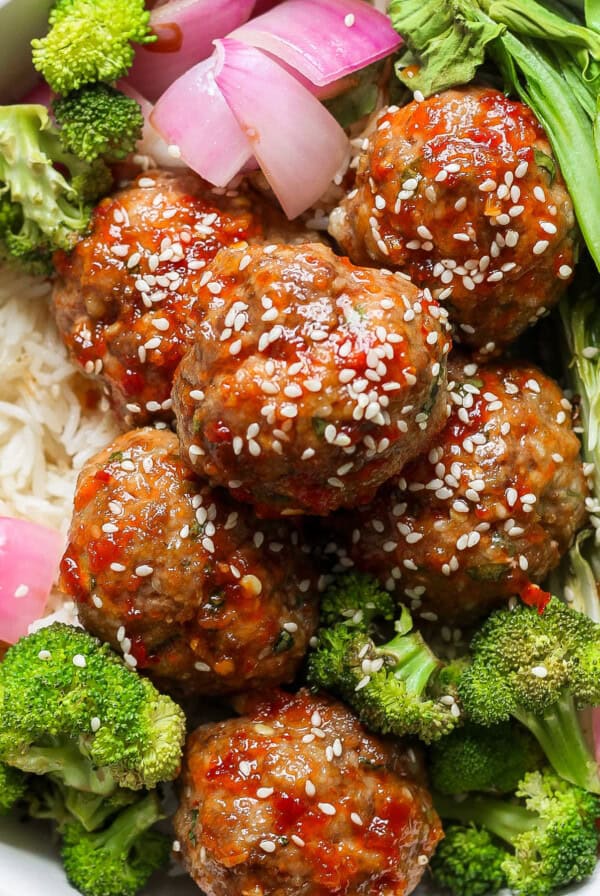 A bowl contains rice, meatballs with sauce and sesame seeds, broccoli, red onion, and bok choy.