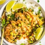 A bowl of skillet-cooked coconut chicken served over white rice, garnished with edamame beans, chopped herbs, and lime wedges.