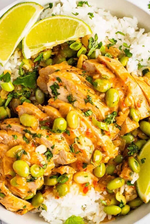 A bowl of skillet-cooked coconut chicken served over white rice, garnished with edamame beans, chopped herbs, and lime wedges.