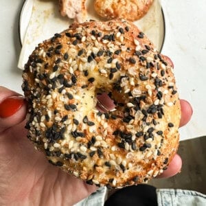 A hand with red nail polish holds an everything bagel with sesame and poppy seeds, with a plate of food blurred in the background.