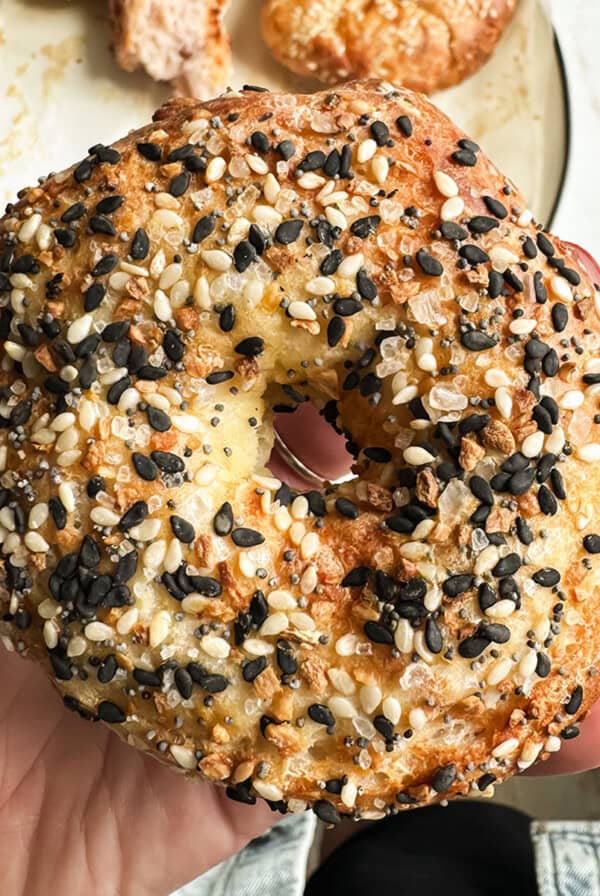 A hand with red nail polish holds an everything bagel with sesame and poppy seeds, with a plate of food blurred in the background.