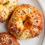 Close-up of three bagels topped with sesame seeds and other seasonings on a beige plate.