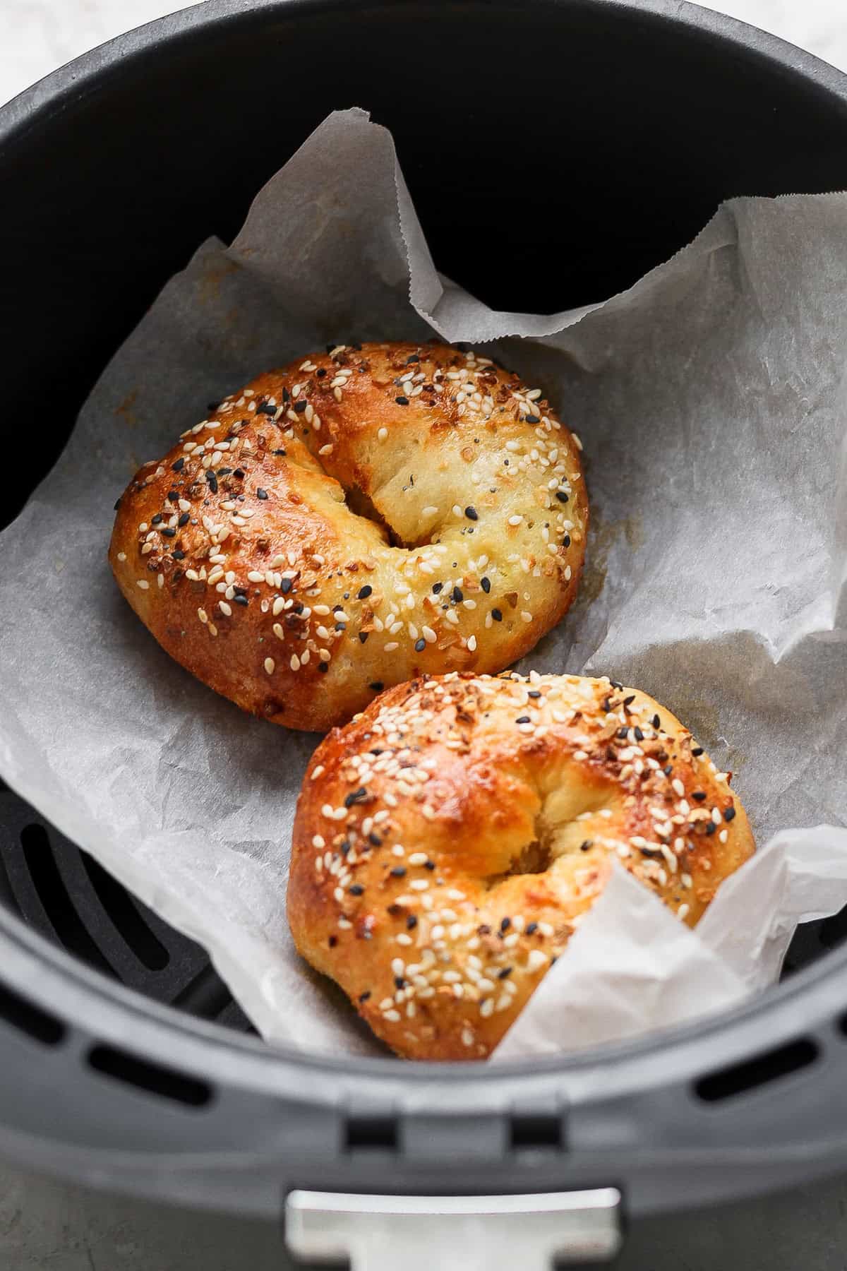 Two bagels topped with sesame and poppy seeds are baking on parchment paper inside an air fryer basket.