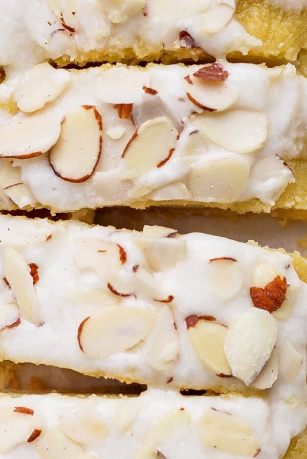 Close-up of almond-topped biscotti with a white icing drizzle. The biscotti pieces are stacked closely together, showcasing the almond slices and the glossy icing.