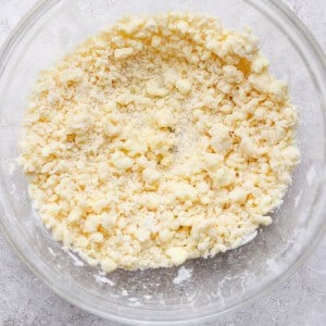 A clear glass bowl containing a crumbly mixture of flour and butter, sitting on a textured gray surface.