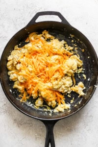 Scrambled eggs with shredded cheese in a black skillet on a light-colored surface.