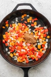 Chopped red, orange, and yellow bell peppers with onions and ground spices in a cast iron skillet.