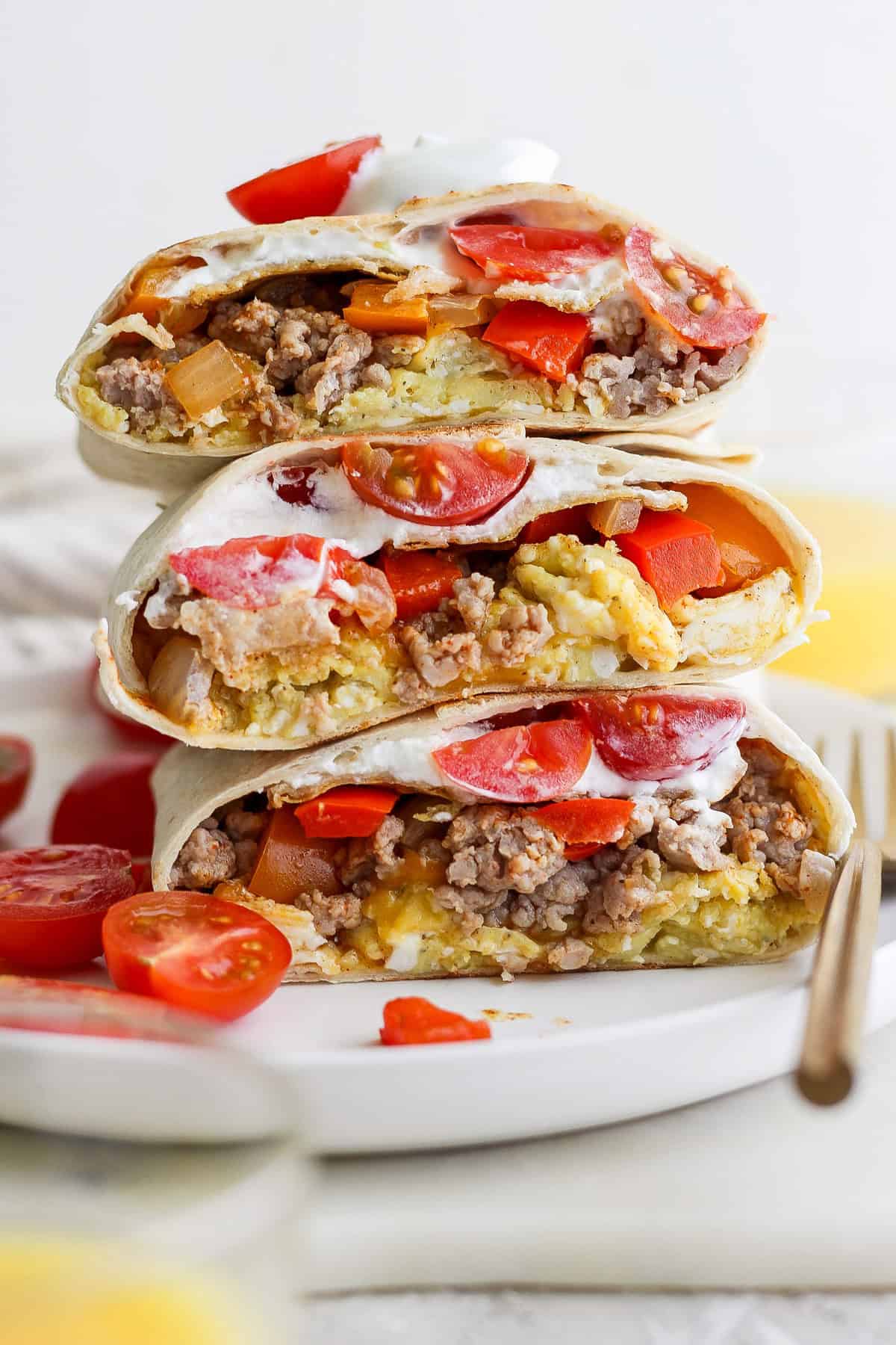 Stacked burritos filled with ground meat, cheese, tomatoes, and sour cream on a white plate. Sliced tomatoes are scattered around.