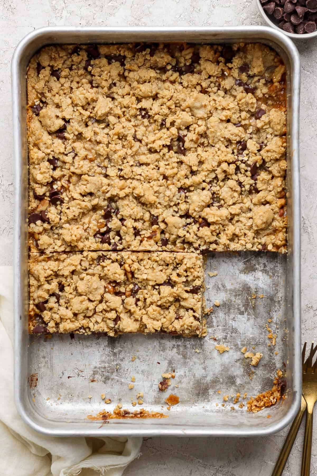 A metal baking pan filled with crumbly dessert bars. Two bars have been cut out, revealing a layered filling, while the rest of the dessert remains uncut. A fork and a napkin are nearby.