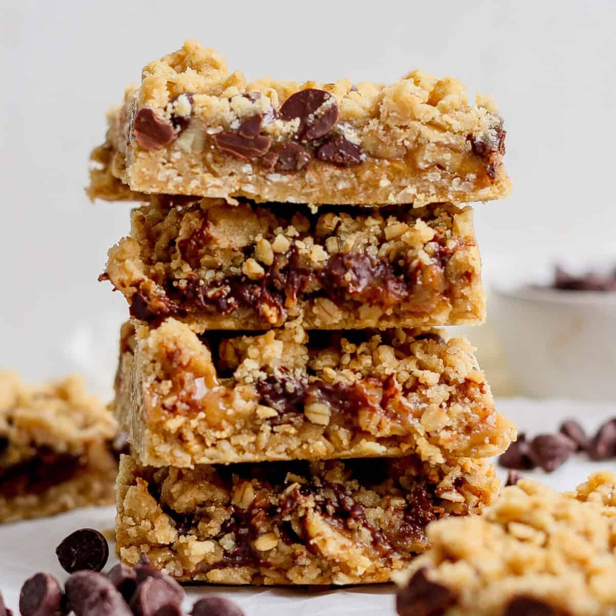 A stack of four chocolate chip oatmeal bars with a crumbly texture is placed on a white surface. Chocolate chips are scattered around the bars.