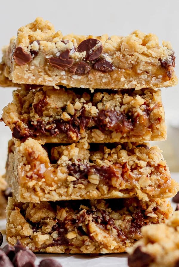 A stack of four chocolate chip oatmeal bars with a crumbly texture is placed on a white surface. Chocolate chips are scattered around the bars.