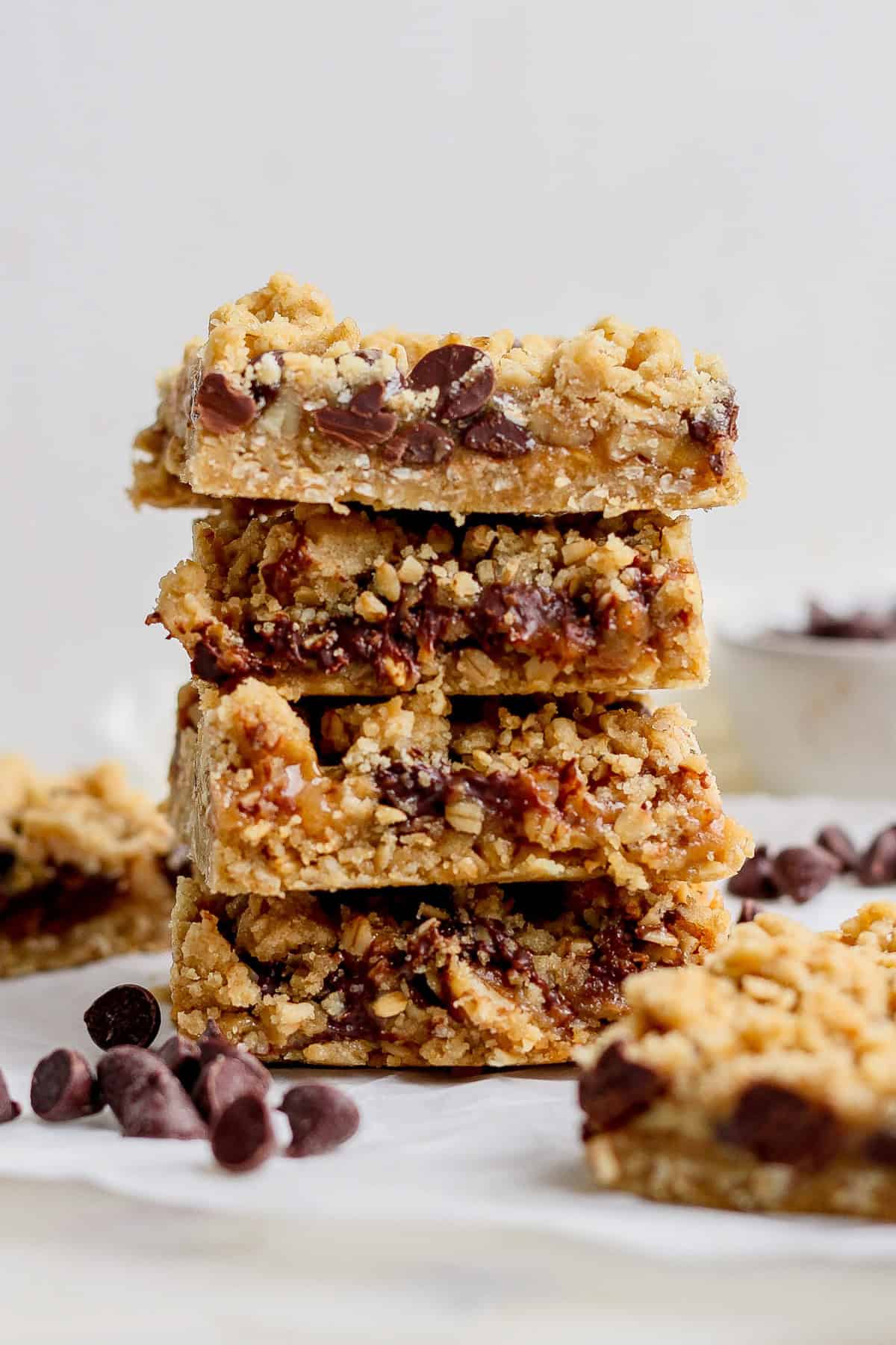 A stack of four chocolate chip oatmeal bars with crumbly tops is displayed on a white surface, surrounded by a few scattered chocolate chips.