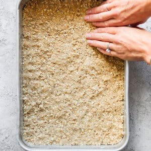 Hands press down a mixture in a square baking pan. The mixture contains visible oats and is evenly spread across the pan.