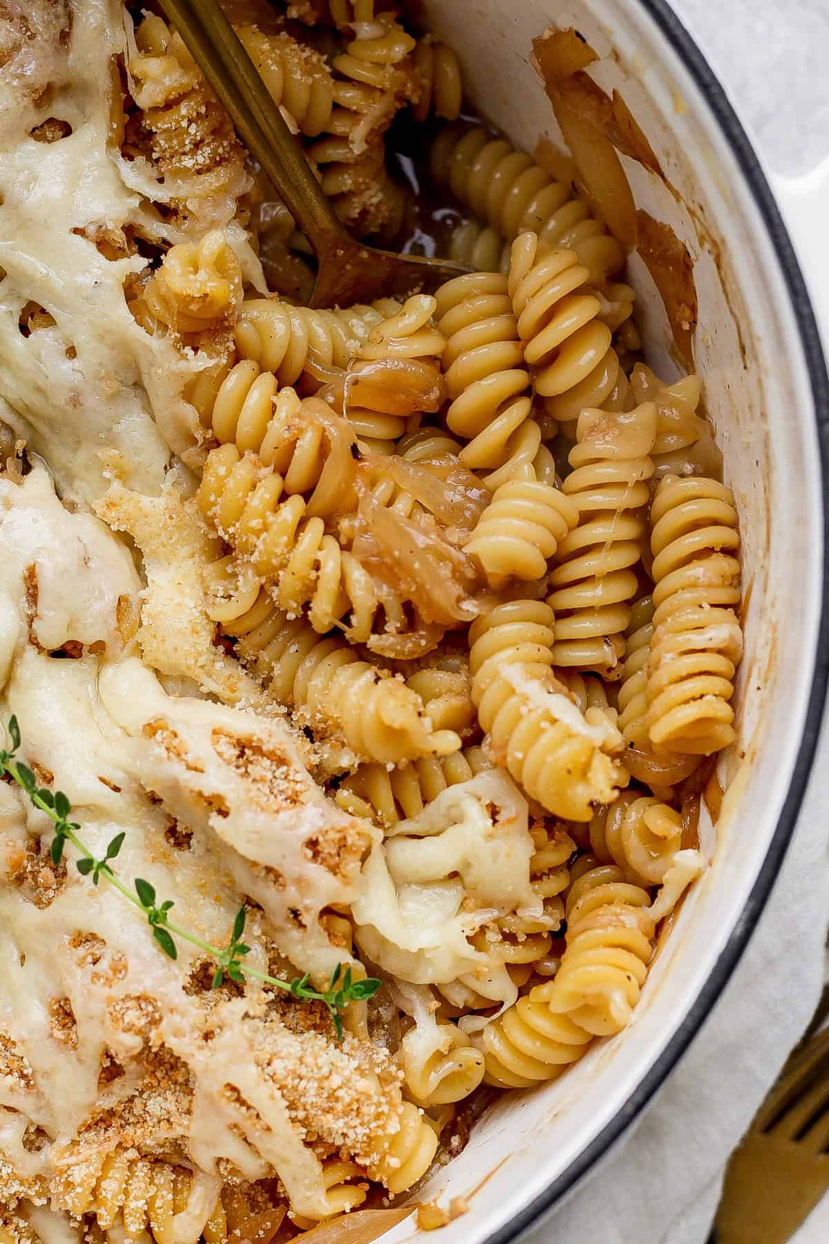 Close-up of baked rotini pasta with melted cheese, breadcrumbs, and herbs in a round dish. A fork is partially visible in the pasta.