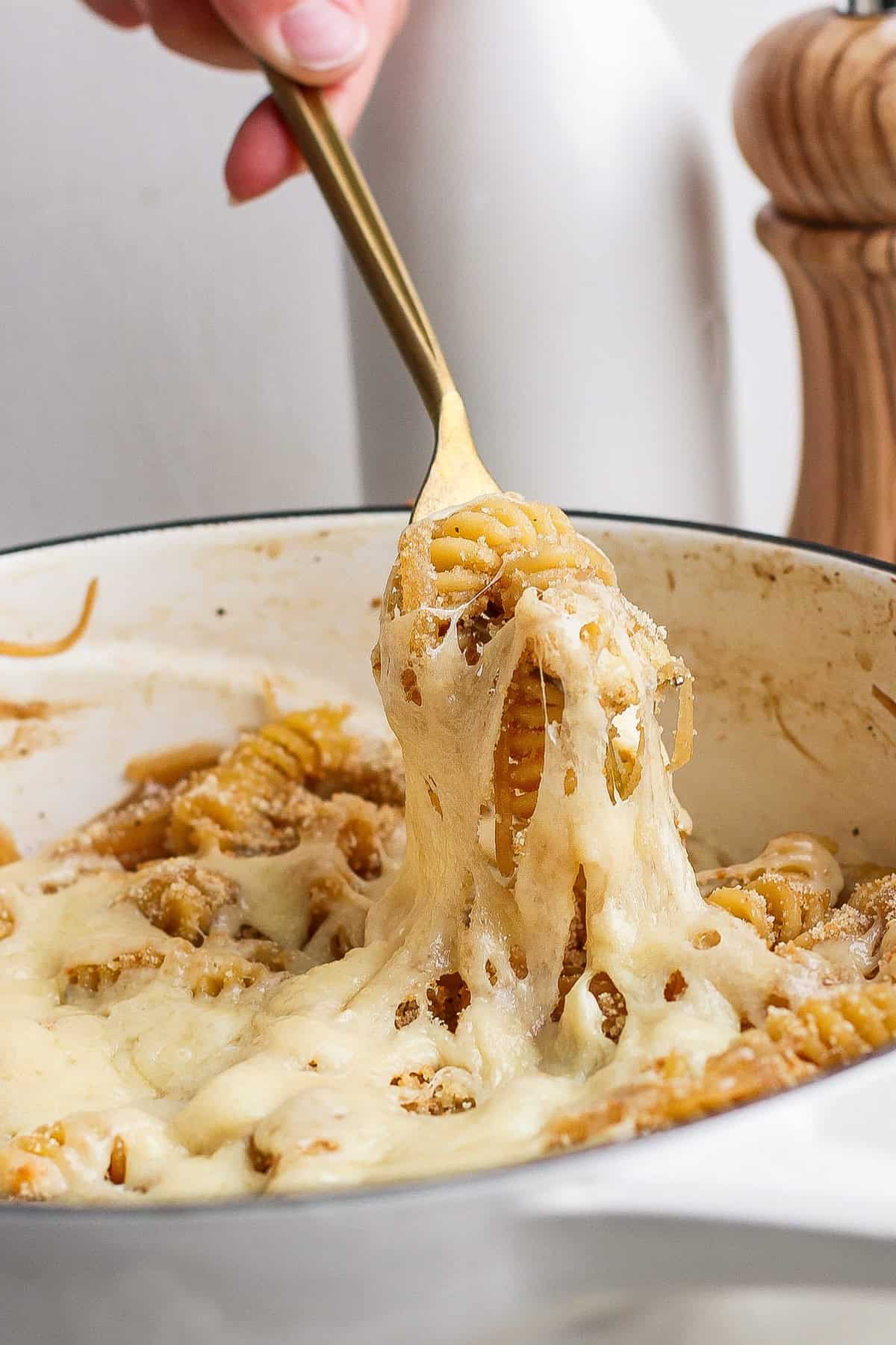 A hand using a golden fork to lift cheesy pasta from a white pot, with melted cheese stretching from the pot.