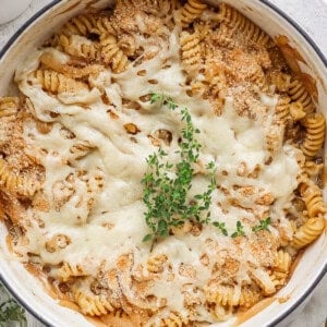 A baked pasta dish in a white pot, featuring rotini pasta covered with melted cheese and garnished with a sprig of fresh herbs on top.