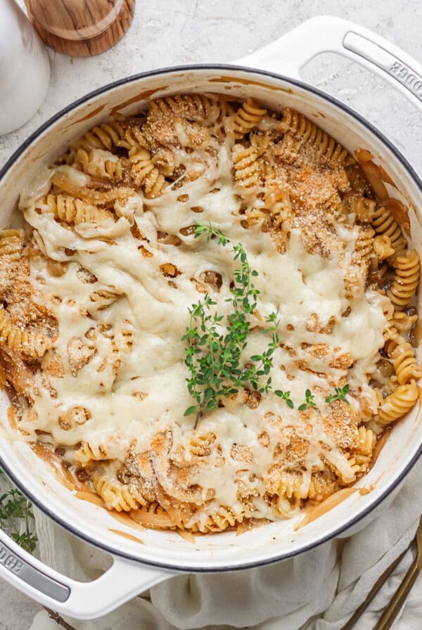 A baked pasta dish in a white pot, featuring rotini pasta covered with melted cheese and garnished with a sprig of fresh herbs on top.