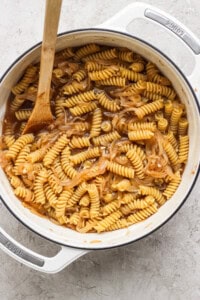 A pot filled with cooked rotini pasta mixed with caramelized onions, with a wooden spoon resting on the side.