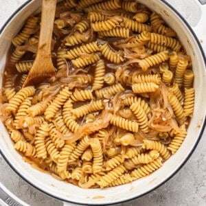 A pot filled with cooked rotini pasta mixed with caramelized onions, with a wooden spoon resting on the side.