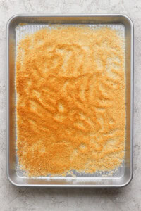 A baking sheet filled with an even layer of finely ground, golden-brown crumbs placed on a light-colored surface.