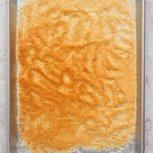 A baking sheet filled with an even layer of finely ground, golden-brown crumbs placed on a light-colored surface.