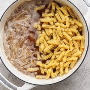 A white pot containing partially cooked rigatoni pasta and bubbling broth, ready to be mixed.