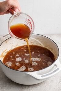 A hand is pouring brown liquid from a Pyrex measuring cup into a white pot filled with similar brown liquid on a kitchen counter.