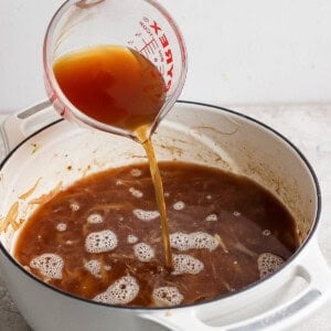 A hand is pouring brown liquid from a Pyrex measuring cup into a white pot filled with similar brown liquid on a kitchen counter.