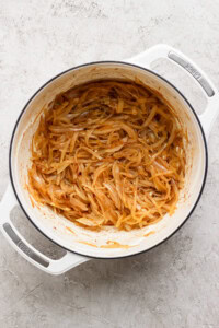 A white pot containing caramelized onions in a light brown sauce on a textured, light-colored surface.