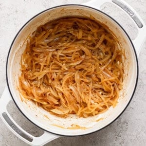 A white pot containing caramelized onions in a light brown sauce on a textured, light-colored surface.