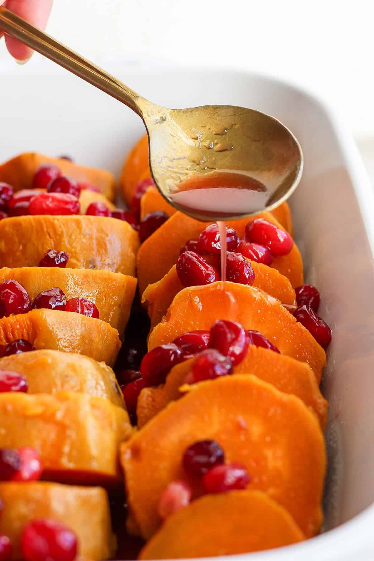 A spoon drizzles liquid over sliced sweet potatoes topped with cranberries in a rectangular white baking dish.