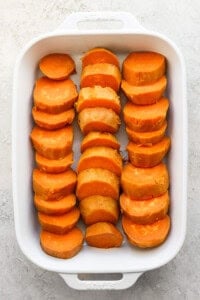 A white baking dish filled with sliced sweet potatoes arranged in three rows on a light surface.