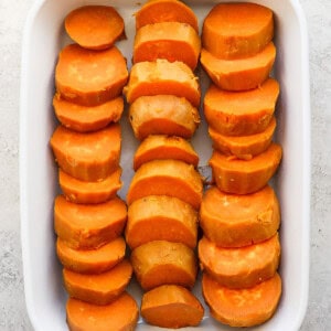 A white baking dish filled with sliced sweet potatoes arranged in three rows on a light surface.