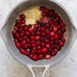 A gray pot contains raw cranberries, a block of butter, and sugar on a light textured surface.