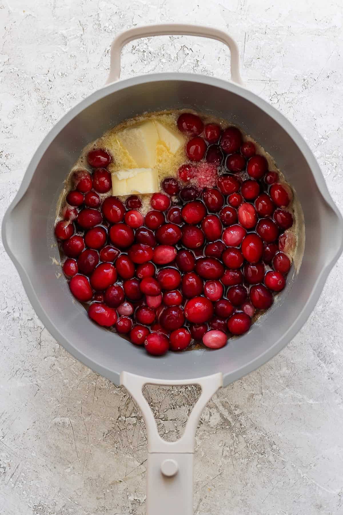 A gray pot contains raw cranberries, a block of butter, and sugar on a light textured surface.