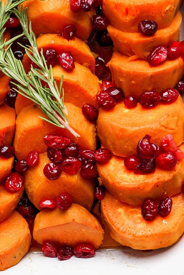 Sliced sweet potatoes topped with cranberries and garnished with a sprig of rosemary, served in a white dish.