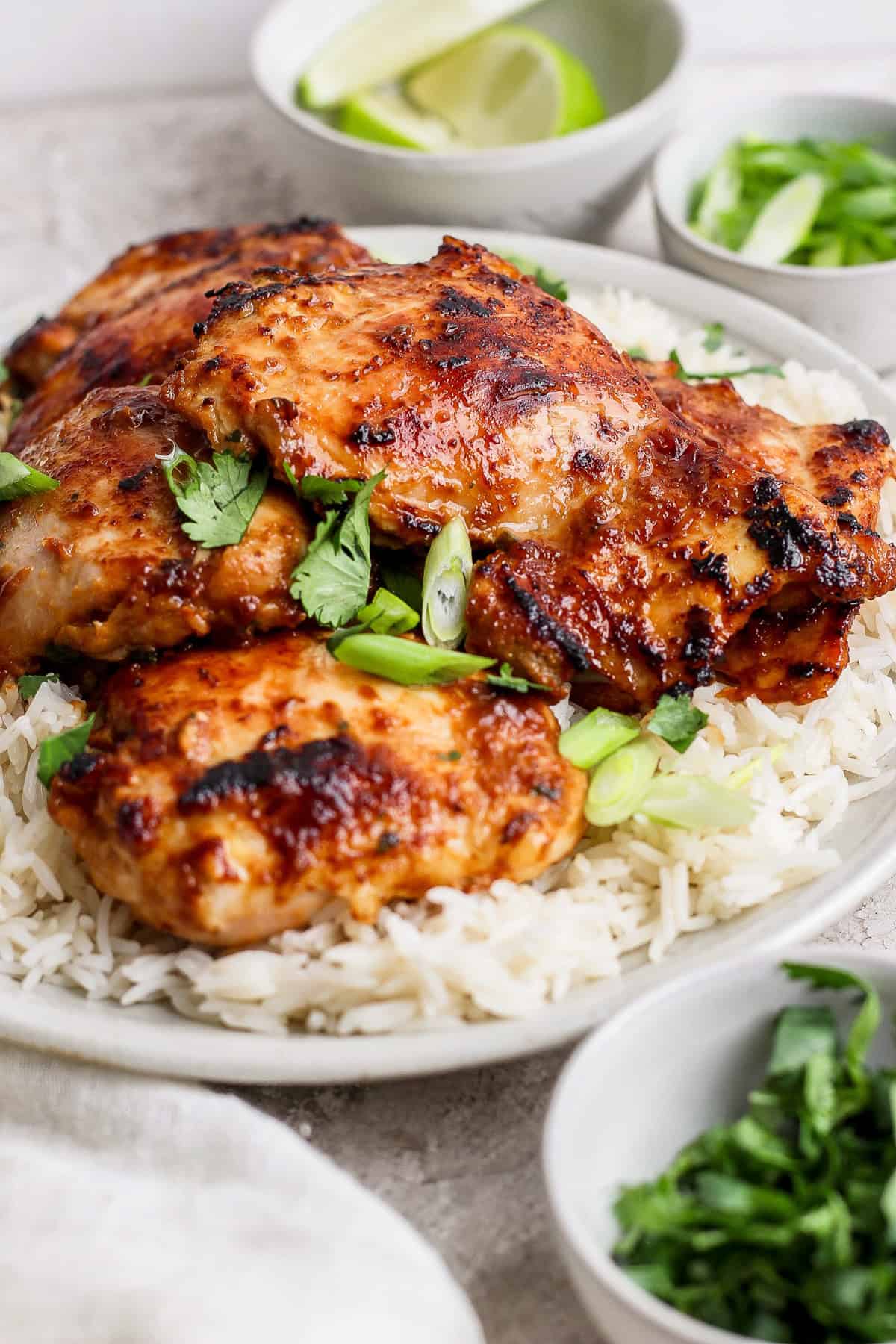 A plate of grilled chicken thighs garnished with herbs on a bed of white rice, accompanied by sliced green onions and lime wedges.