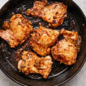 A cast-iron skillet contains five pieces of cooked, seasoned chicken, set on a light-colored surface.