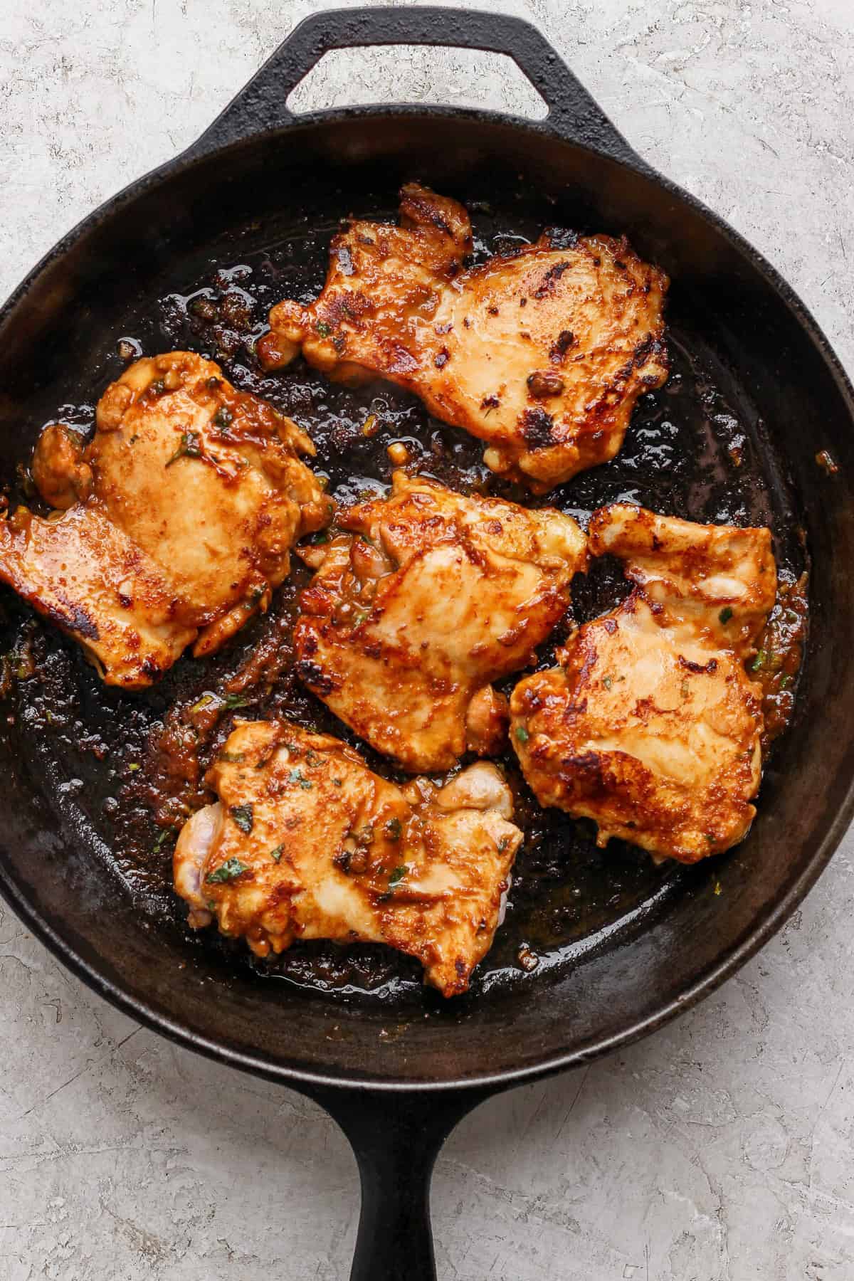 A cast-iron skillet contains five pieces of cooked, seasoned chicken, set on a light-colored surface.