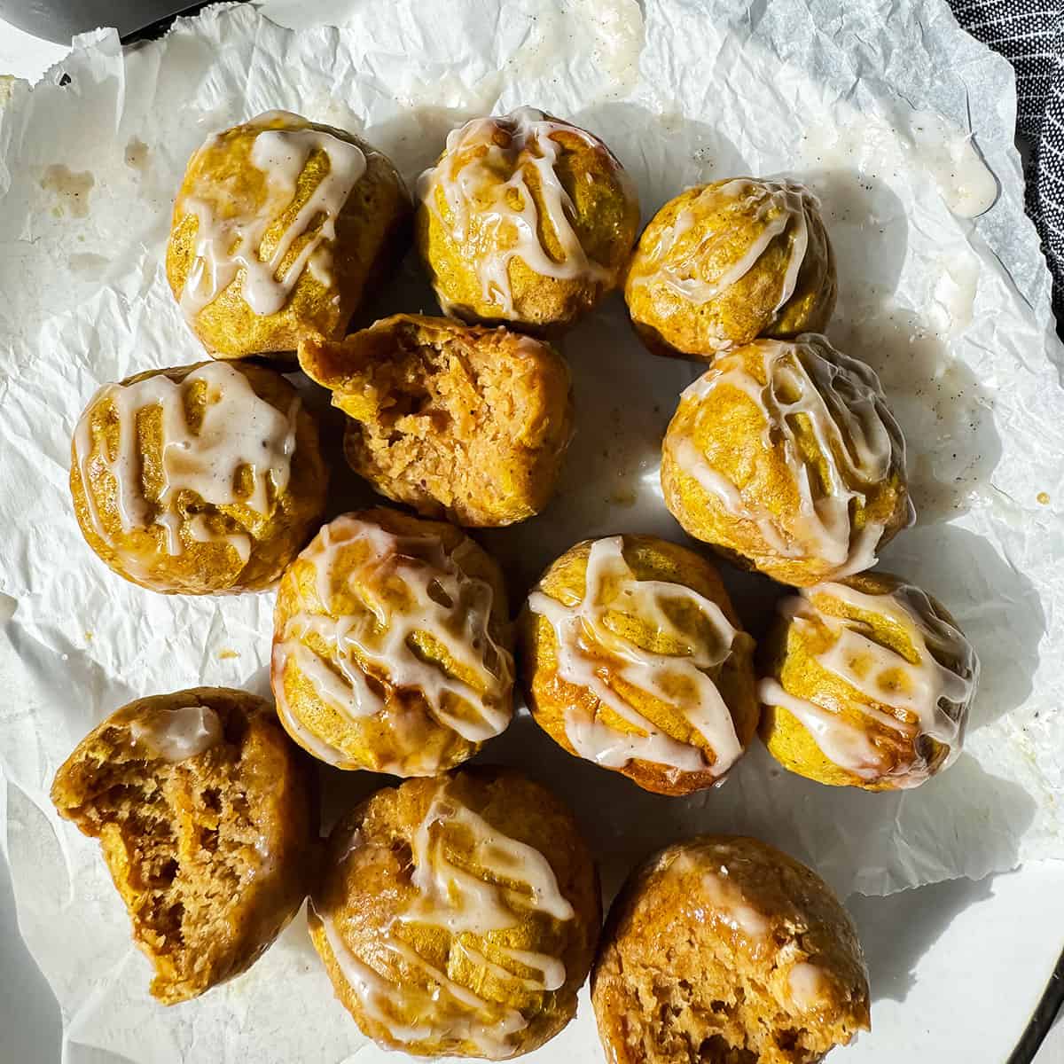 A dozen small, round cakes with white icing drizzle, some with bites taken out, are arranged on crumpled parchment paper under natural light.