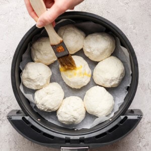 A hand brushes egg wash on dough balls arranged in an air fryer basket lined with parchment paper.