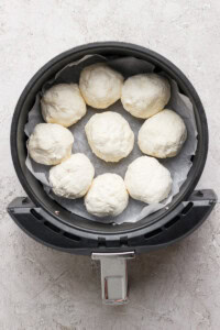 An air fryer basket lined with parchment paper holds eight uncooked dough balls in a circular arrangement.