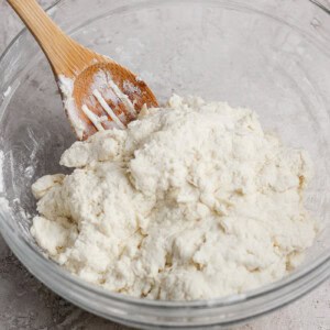 A wooden spoon rests in a glass bowl filled with a mixture of flour and a white ingredient, likely dough, on a textured countertop.