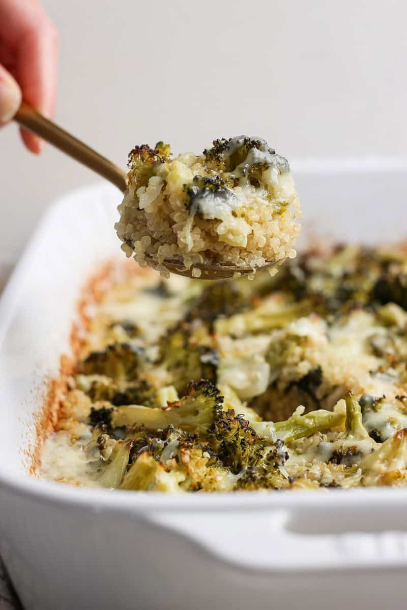 A spoon scooping a portion of baked quinoa and broccoli from a white dish.