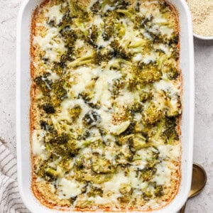 A baked casserole in a white dish with melted cheese and broccoli, accompanied by bowls of grated cheese and breadcrumbs. A striped towel and two spoons are nearby.