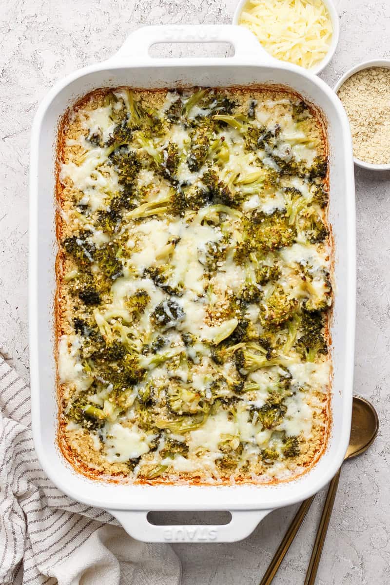 Baked broccoli casserole with melted cheese in a white dish, next to bowls of grated cheese and breadcrumbs on a textured surface.