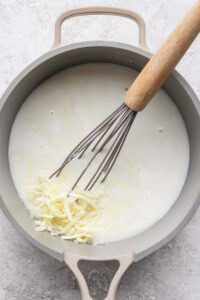 A pot containing a creamy liquid mixture with shredded cheese being whisked in.