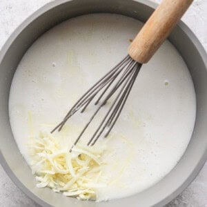 A pot containing a creamy liquid mixture with shredded cheese being whisked in.